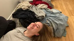 Donna sorridente sdraiata accanto a una pila di vestiti sul pavimento di legno.
