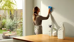 Donna pulisce una parete della cucina con prodotti per la pulizia vicino a una finestra luminosa.