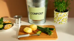 Patate sbucciate e ortaggi su tagliere, secchio compost, erbe aromatiche in cucina.