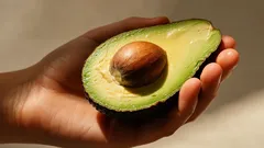 Mano che tiene mezzo avocado maturo con nocciolo, su sfondo neutro, luce naturale.